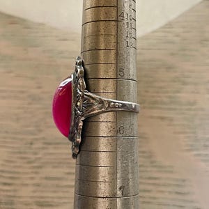May include: A silver ring with a large, oval, fuchsia-colored stone. The ring has an ornate, filigree design around the stone. The ring is being measured on a metal ring sizer. The ring size appears to be between a 5 and a 6.