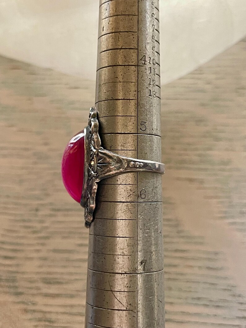 May include: A silver ring with a large, oval, fuchsia-colored stone. The ring has an ornate, filigree design around the stone. The ring is being measured on a metal ring sizer. The ring size appears to be between a 5 and a 6.