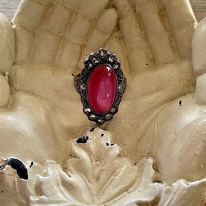 May include: An antique-style ring with a large oval, pink gemstone set in a dark metal frame. The frame is detailed with small, silver-colored accents. The ring is displayed on a cream-colored, hand-shaped decorative dish.