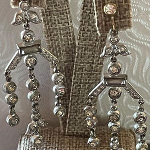 May include: Silver-toned chandelier earrings with multiple dangling strands of clear, round gemstones. The earrings feature decorative floral and geometric elements, creating a vintage-inspired design. The earrings are displayed on a textured, neutral-colored surface.