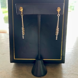May include: Gold-toned dangle earrings with a post backing. The earrings feature a small, round stud and a chain of small, gold and silver-colored beads. The earrings are displayed on a black jewelry stand.