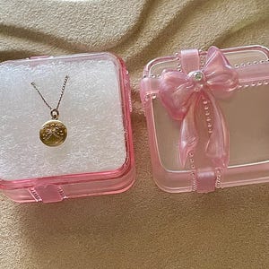 May include: A gold-toned necklace with a circular pendant featuring an embossed angel design, resting inside a square, translucent pink plastic box. The box lid is decorated with a molded pink bow and a small, clear embedded rhinestone.