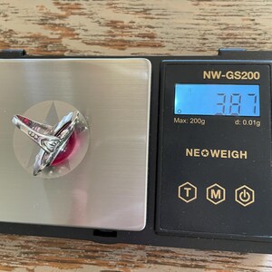 May include: A silver ring with a large, round, red gemstone is displayed on a digital scale. The scale reads 3.87 grams. The ring has an ornate, detailed design around the gemstone.