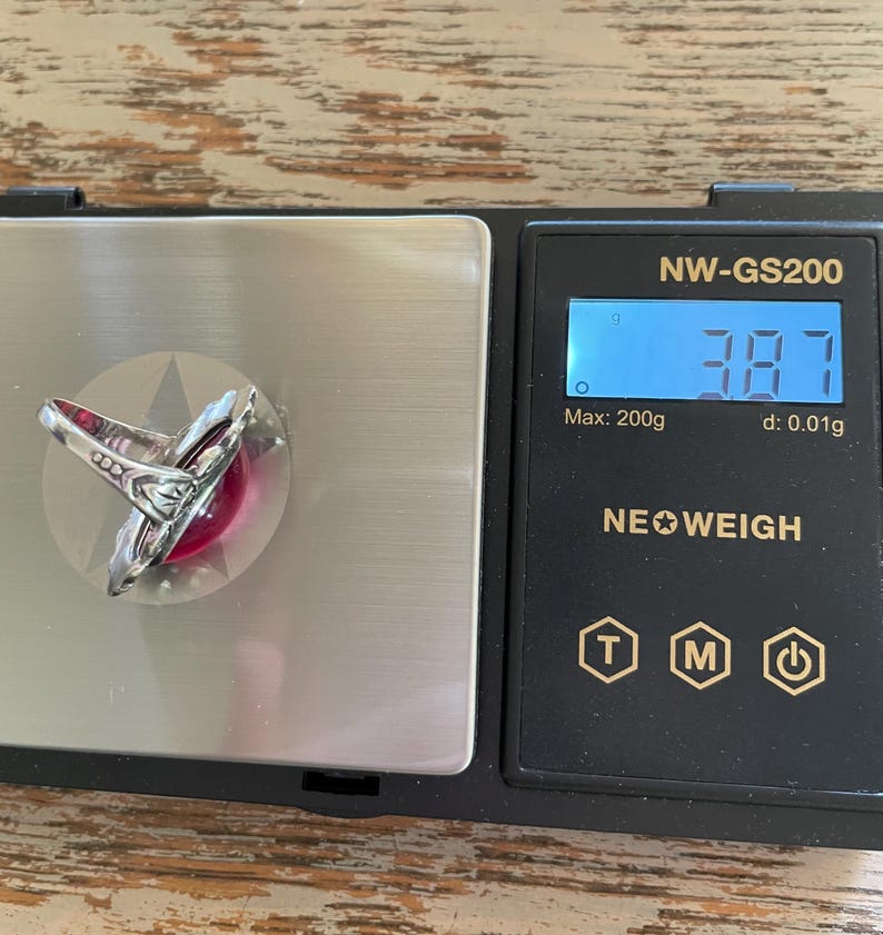 May include: A silver ring with a large, round, red gemstone is displayed on a digital scale. The scale reads 3.87 grams. The ring has an ornate, detailed design around the gemstone.