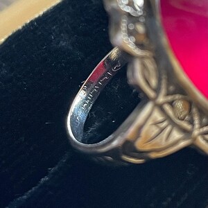 May include: Close-up of a silver ring with a large, oval, fuchsia-colored stone. The ring has an ornate, floral design on the band and around the stone. The inside of the band has stamped markings. The ring is set against a dark background.