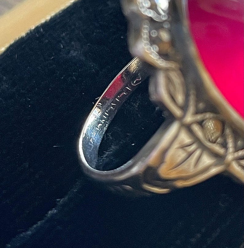May include: Close-up of a silver ring with a large, oval, fuchsia-colored stone. The ring has an ornate, floral design on the band and around the stone. The inside of the band has stamped markings. The ring is set against a dark background.