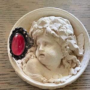 May include: A silver ring with a large oval, pink gemstone is displayed on a white, circular relief sculpture of an angel's face. The ring has a dark, ornate setting with small, round accents.