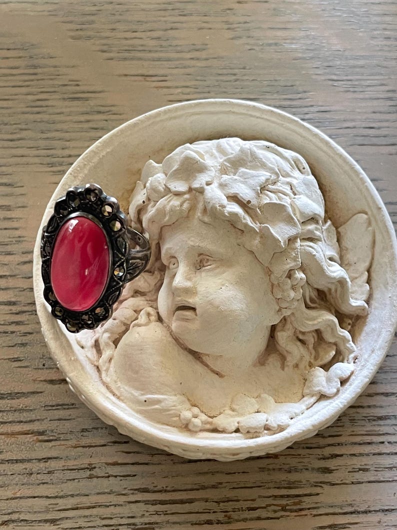 May include: A silver ring with a large oval, pink gemstone is displayed on a white, circular relief sculpture of an angel's face. The ring has a dark, ornate setting with small, round accents.