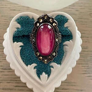 May include: A silver-toned ring with a large, oval, pink gemstone. The ring is set in a decorative frame with small, round accents. The ring rests on a teal fabric inside a white, heart-shaped jewelry box with ornate edges.
