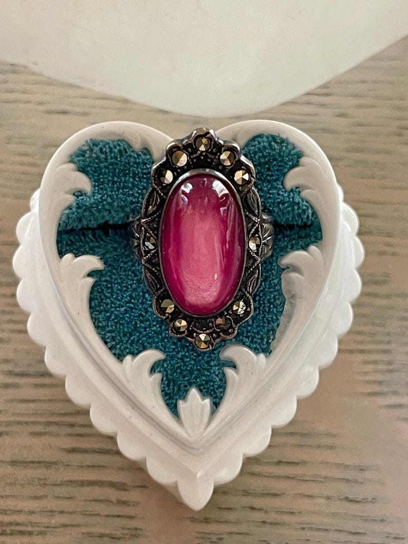 May include: A silver-toned ring with a large, oval, pink gemstone. The ring is set in a decorative frame with small, round accents. The ring rests on a teal fabric inside a white, heart-shaped jewelry box with ornate edges.