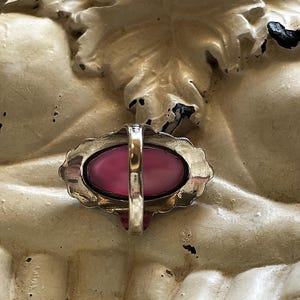 May include: A vintage ring with a large, oval, pink gemstone set in a gold-toned metal frame. The ring has a decorative, scalloped edge and a simple band. The ring is set against a textured, off-white background.