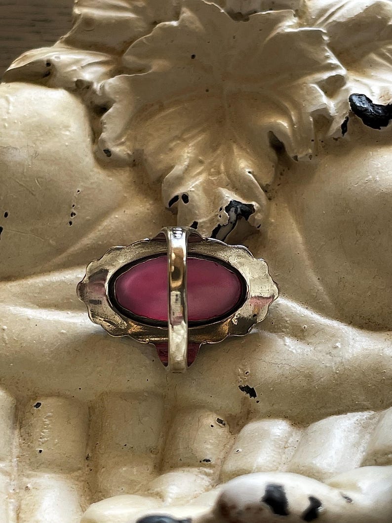 May include: A vintage ring with a large, oval, pink gemstone set in a gold-toned metal frame. The ring has a decorative, scalloped edge and a simple band. The ring is set against a textured, off-white background.