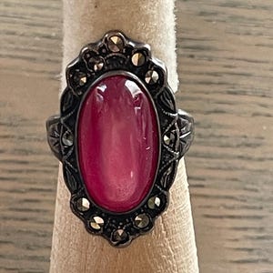 May include: A vintage silver ring featuring an oval, cabochon-cut, pink gemstone. The ring has an ornate, dark metal setting with decorative accents and small, sparkling stones. The ring is displayed on a neutral-colored ring holder.