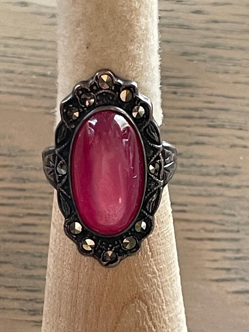 May include: A vintage silver ring featuring an oval, cabochon-cut, pink gemstone. The ring has an ornate, dark metal setting with decorative accents and small, sparkling stones. The ring is displayed on a neutral-colored ring holder.