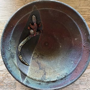 May include: A handmade, dark brown ceramic bowl with a textured surface and a decorative lizard figurine. The bowl has a unique color gradient, transitioning from dark brown to shades of blue and red.