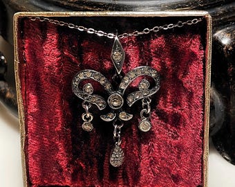 May include: A silver-toned metal pendant necklace featuring a fleur-de-lis shape encrusted with small clear stones. The pendant hangs from a delicate chain and includes three dangling charms, with a central teardrop-shaped drop at the bottom.
