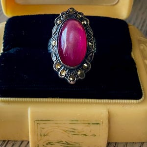 May include: A vintage silver ring with a large oval, magenta-colored stone. The ring is set in an ornate, filigree-style setting with small, gold-colored accents. The ring is displayed in a yellow jewelry box with a black velvet interior.