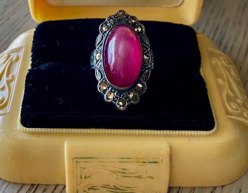 May include: A vintage silver ring with a large oval, magenta-colored stone. The ring is set in an ornate, filigree-style setting with small, gold-colored accents. The ring is displayed in a yellow jewelry box with a black velvet interior.