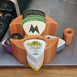 May include: A 3D-printed orange guitar pick holder with multiple compartments, filled with various guitar picks. The picks are in different colors and designs, including a white pick with a black 'M' and a green pick with text.