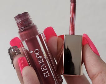May include: A clear glass bottle with a rose-colored liquid inside. The bottle has a gold cap and the word "ELEVISPO" printed on the side. The bottle is being held by a hand with pink nail polish.