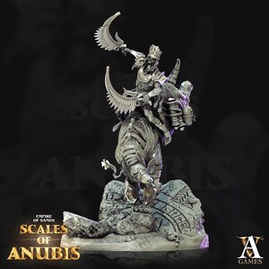 May include: A detailed miniature figurine of a skeletal figure riding a winged, skeletal beast. The figure is holding a scimitar and wears a crown and jewelry. The base is a rocky outcropping with hieroglyphics. The text "Empire of Sands Scales of Anubis" is visible on the base.