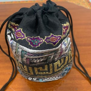 May include: A small, black drawstring bag with intricate embroidery. The bag features a detailed pattern in gold, purple, and green. The top of the bag is black, and the drawstring is a black cord.