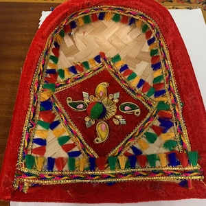 May include: A decorative, fan-shaped item with a red velvet border. The center features woven bamboo and a red square with gold embroidery and colorful tassels. The design includes a diamond shape and paisley motifs.