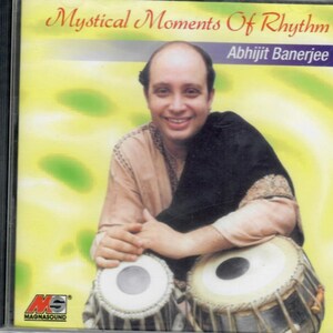 Może przedstawiać: Pudełko na płytę CD z tytułem "Mystical Moments Of Rhythm" i nazwiskiem "Abhijit Banerjee". Obraz przedstawia uśmiechniętego mężczyznę siedzącego z dwoma bębnami tabla. Tło jest żółte z rozmytym wzorem. Widoczne jest logo MagnaSound.
