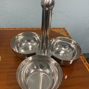 May include: A stainless steel condiment server with three round bowls and a central handle topped with a sphere. The bowls are arranged in a triangular pattern, reflecting light.