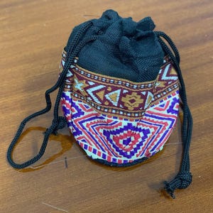 May include: A small drawstring pouch with a black top and a patterned bottom. The bottom features a geometric design in red, purple, white, and brown. The drawstring is black.