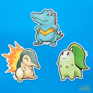 Op de afbeelding: Drie cartoon stickers met Pokémon-personages. De eerste sticker is een blauwe en gele Feraligatr, de tweede is een bruine en oranje Cyndaquil en de derde is een groene en bruine Bayleef.