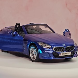 Puede incluir: Modelo a escala azul de un BMW Z4 descapotable con las puertas abiertas. El coche de juguete tiene ruedas plateadas e interior negro. La matrícula dice "CHARLIE". El coche está sobre una superficie blanca con fondo rosa. El logo "CarModels Garage" está arriba a la izquierda.
