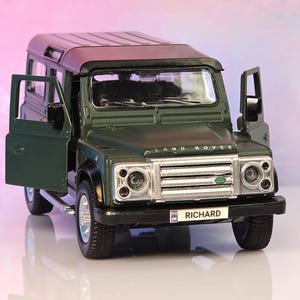 May include: A dark green Land Rover toy vehicle with open front doors. The front grille features the Land Rover logo, and the license plate displays the name RICHARD with a small UK flag icon. The model has black trim, silver wheels, and clear windows.
