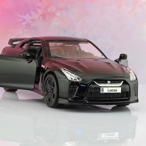 May include: A black die-cast model sports car with an open driver-side door. The front number plate features the name Lucas with a small flag icon. The vehicle has dark wheels, a rear spoiler, and clear headlights, reflecting on a white surface.