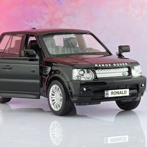 May include: A black Range Rover toy car model with an open driver-side door. The vehicle features silver wheels, clear headlights, and a front number plate that reads RONALD with a small UK flag icon. The bonnet displays the text RANGE ROVER.
