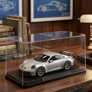 May include: A silver diecast model of a Porsche 911 GT3 sports car enclosed in a clear rectangular acrylic display case with a black base. The model features detailed wheels, a rear spoiler, and side mirrors, resting on a flat surface.