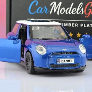 Könnte beinhalten: Ein blau-weißes Modellauto mit geöffneten Türen. Das vordere Nummernschild trägt die Aufschrift DANIEL mit einer kleinen britischen Flagge. Das Auto hat schwarze Räder, ein weißes Dach und klare Scheinwerfer. Im Hintergrund eine schwarze Box mit Car Models Garage.