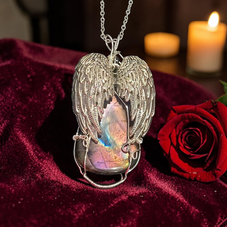May include: A silver-toned pendant necklace featuring a large, iridescent gemstone encased in wire, with detailed angel wing accents. The gemstone displays hues of purple, blue, and green. The necklace is displayed on a deep red velvet surface.