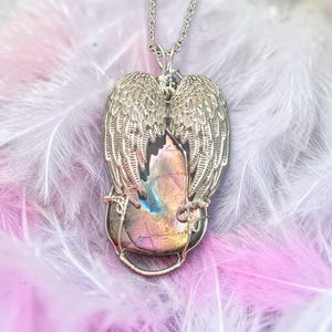 May include: Silver-toned pendant necklace featuring a large, iridescent gemstone encased in a wire frame. The gemstone is flanked by detailed, openwork angel wings. The necklace chain is also silver-toned, creating a cohesive and elegant design.