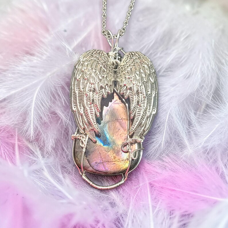 May include: Silver-toned pendant necklace featuring a large, iridescent gemstone encased in a wire frame. The gemstone is flanked by detailed, openwork angel wings. The necklace chain is also silver-toned, creating a cohesive and elegant design.