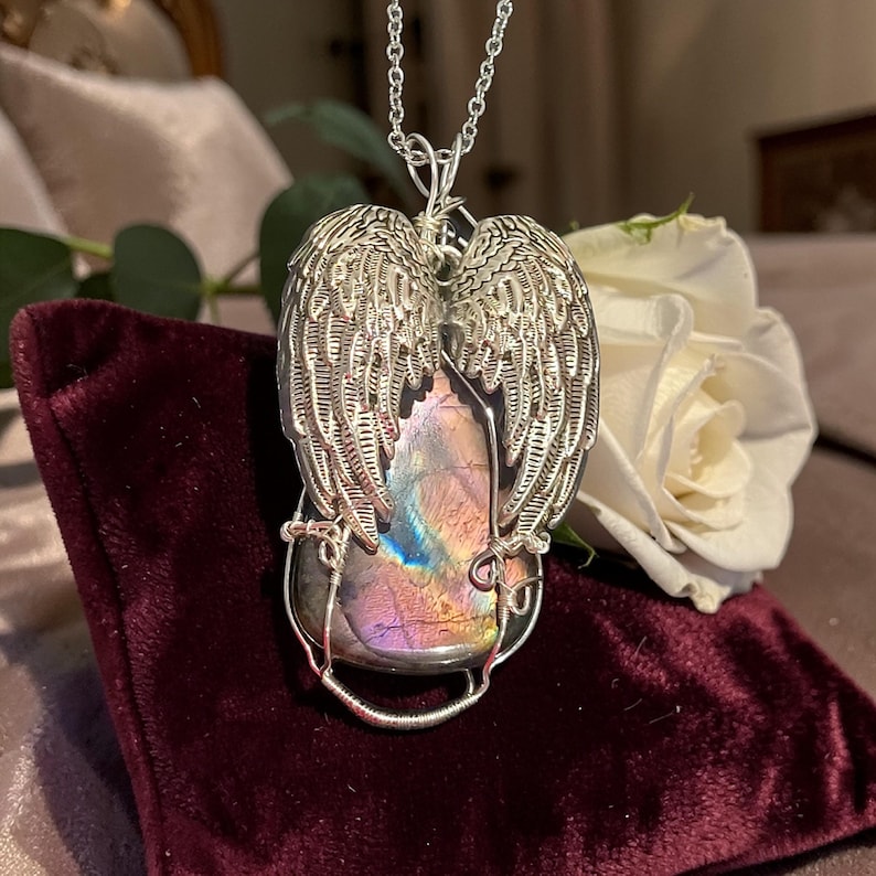 May include: A silver-toned pendant necklace featuring a large, iridescent gemstone framed by detailed angel wings. The gemstone displays hues of purple, pink, and blue. The pendant hangs from a delicate chain, showcasing a unique, handcrafted jewelry design.