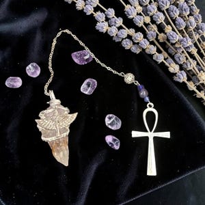 Puede incluir: Un péndulo de tono plateado con un cristal transparente y puntiagudo envuelto en alambre con una figura alada de metal. La cadena incluye una cuenta texturizada, una piedra azul oscuro y un dije de ankh plateado, rodeado de piedras de amatista.