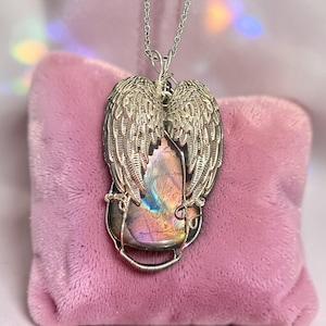 May include: A silver-toned pendant necklace featuring a large, iridescent gemstone framed by detailed angel wings. The gemstone displays a range of colors, including purple, blue, and gold. The necklace is displayed on a pink velvet surface.
