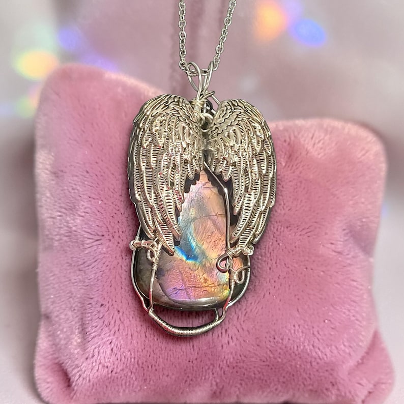 May include: A silver-toned pendant necklace featuring a large, iridescent gemstone framed by detailed angel wings. The gemstone displays a range of colors, including purple, blue, and gold. The necklace is displayed on a pink velvet surface.