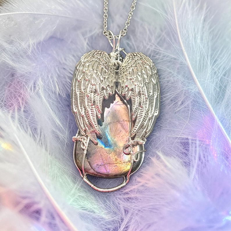 May include: A silver-toned pendant necklace featuring a central, iridescent stone cradled by detailed angel wings. The stone displays hues of purple, blue, and gold. The wings are intricately designed, and the necklace chain is delicate.