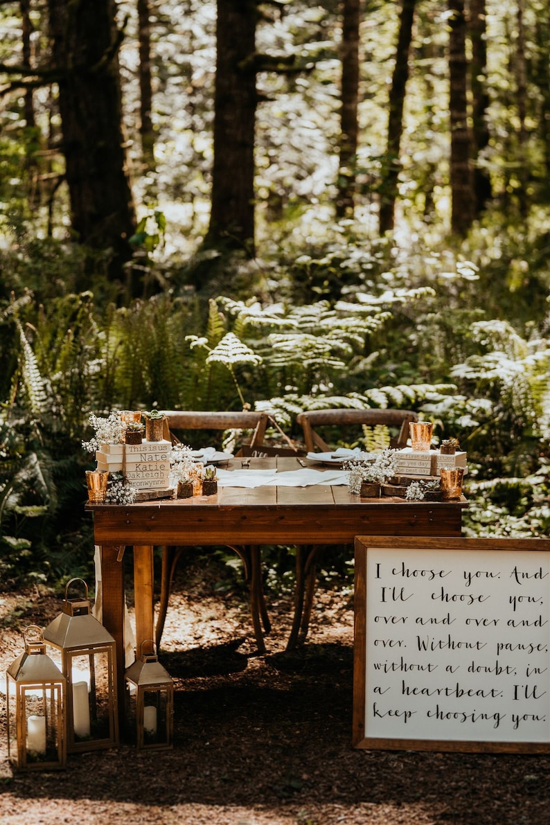 K&ouml;nnte beinhalten: Ein Holztisch f&uuml;r zwei Personen, der in einer Waldlandschaft gedeckt ist. Der Tisch ist mit Gr&uuml;npflanzen, Kerzen und einem Schild dekoriert, auf dem steht: "Ich w&auml;hle dich. Und ich werde dich w&auml;hlen, immer und immer wieder. Ohne Pause, ohne Zweifel, im Herzensschlag. Ich werde dich immer wieder w&auml;hlen."