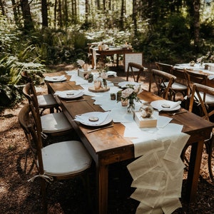May include: A rustic wooden table set with white linens and a book page runner. The table is decorated with flowers, candles, and other centerpieces. The table is set in a forest setting with trees in the background.