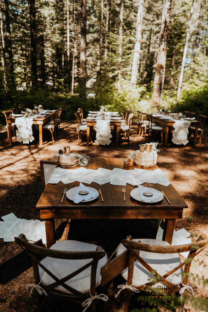 K&ouml;nnte beinhalten: Eine rustikale Hochzeitsempfangstisch-Dekoration in einem Wald mit zwei Gedecken, jedes mit einem wei&szlig;en Teller, goldenem Besteck und einem Holz-Platzkarte, auf dem "Bride" steht.