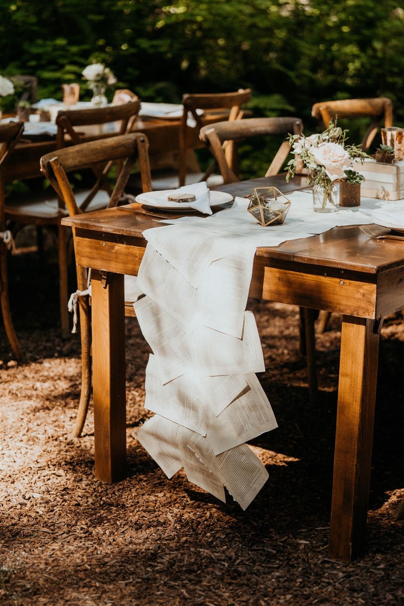K&ouml;nnte beinhalten: Ein rustikaler Holztisch mit einer wei&szlig;en Tischdecke und einem Tischl&auml;ufer aus Buchseiten. Der Tisch ist mit einem Teller, einer Serviette und einer kleinen Glasvase mit Blumen gedeckt. In der Mitte des Tisches befindet sich ein geometrisches Glasornament.