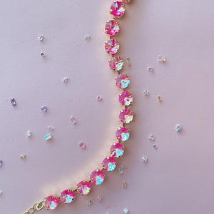 Könnte beinhalten: Ein goldfarbenes Armband mit einer Reihe von schillernden, rosa Edelsteinen. Die runden Edelsteine sind eng aneinander gesetzt. Der Hintergrund ist ein zartes Rosa, mit verstreuten schillernden Kristallen.